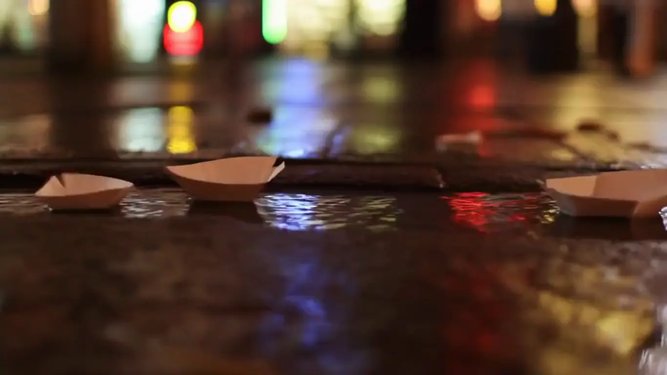 Paper boats drifting down a rain-fed gutter stream past reflected shop lights, cinematic warm-neutral color grade, soft directional key light, 35mm lens, shallow depth of field, natural skin tones, subtle film grain
